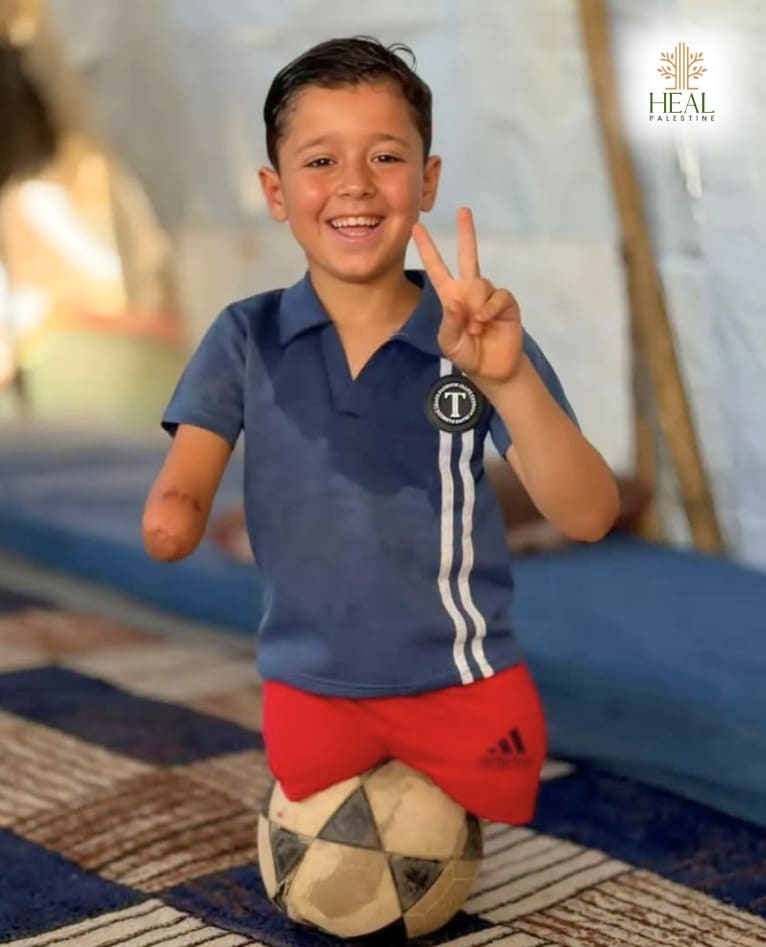 A smiling boy in Gaza flashing a peace sign while kneeling on a football