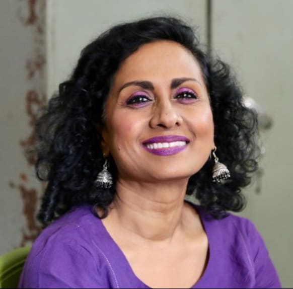 Headshot of Chitra, a woman with brown skin and dark brown hair, who is wearing a purple shirt with matching purple lipstick and eyeshadow.