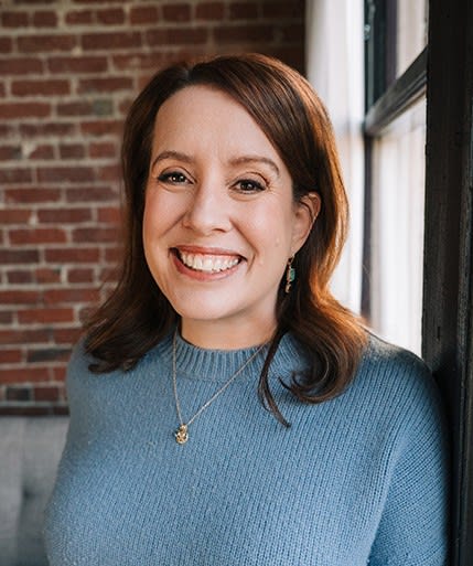 Headshot of Julia Quinn, a smiling woman with brunette hair, wearing a light blue sweater and in front of a brick wall.